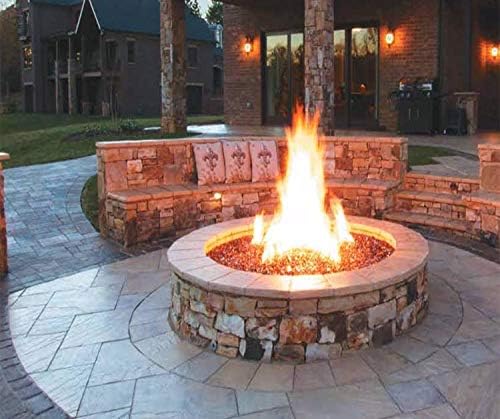 A 19 Firegear Match Light Gas Fire Pit Burner Kit creates a lively flame in a cozy outdoor fire pit, surrounded by circular stone seating on a patio. In front of a modern house with stone and brick accents, soft outdoor lighting enhances the ambiance.